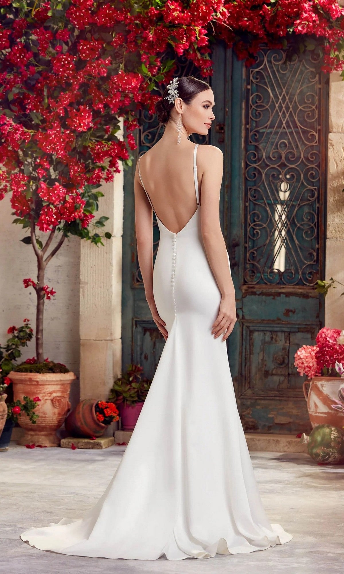 A woman in the Long White Dress: Alyce 7051 with a cowl neckline stands outdoors by a rustic green door, surrounded by red flowers and potted plants.